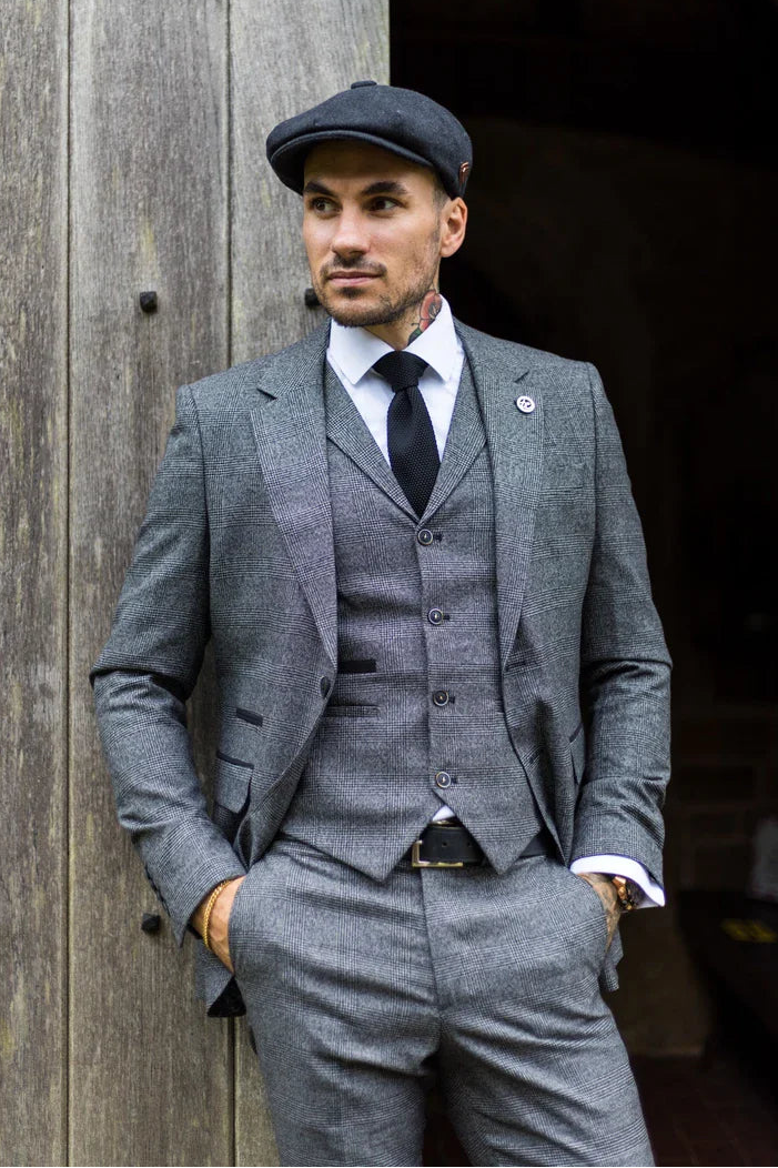 Άντρας με γκρι κοστούμι και καπέλο στέκεται δίπλα σε έναν ξύλινο τοίχο.